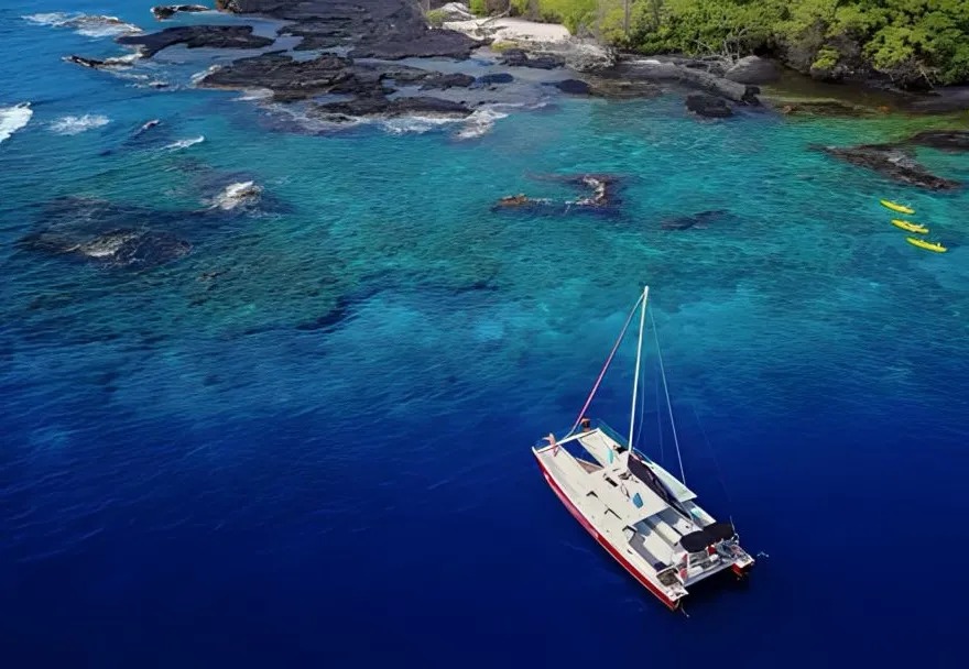 Boat on deep blue water near rocky shoreline and green trees, with kayaks visible in clear water.