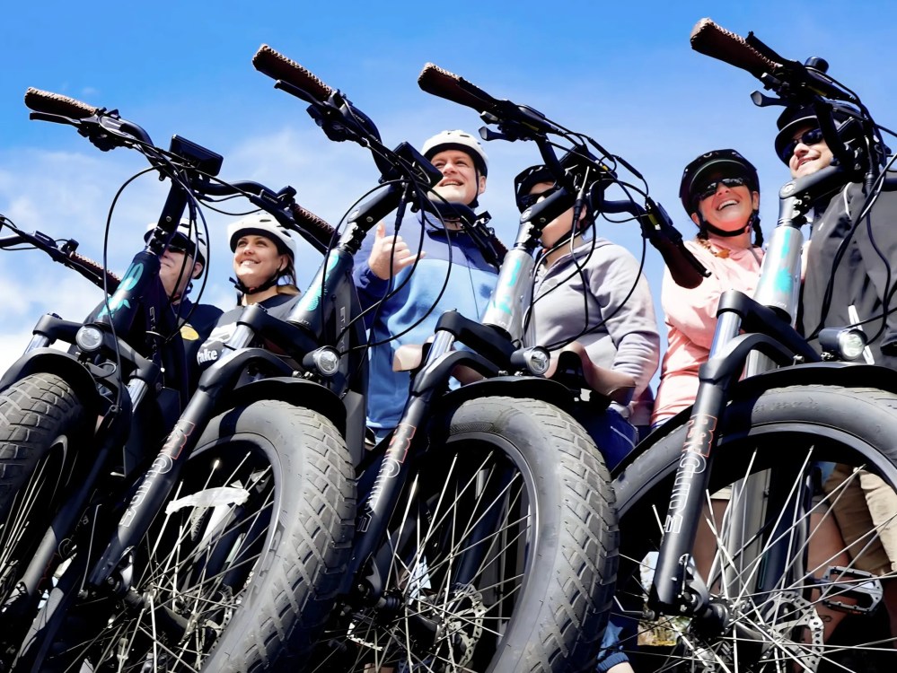 Group of people with helmets standing behind bicycles, smiling against a clear blue sky.