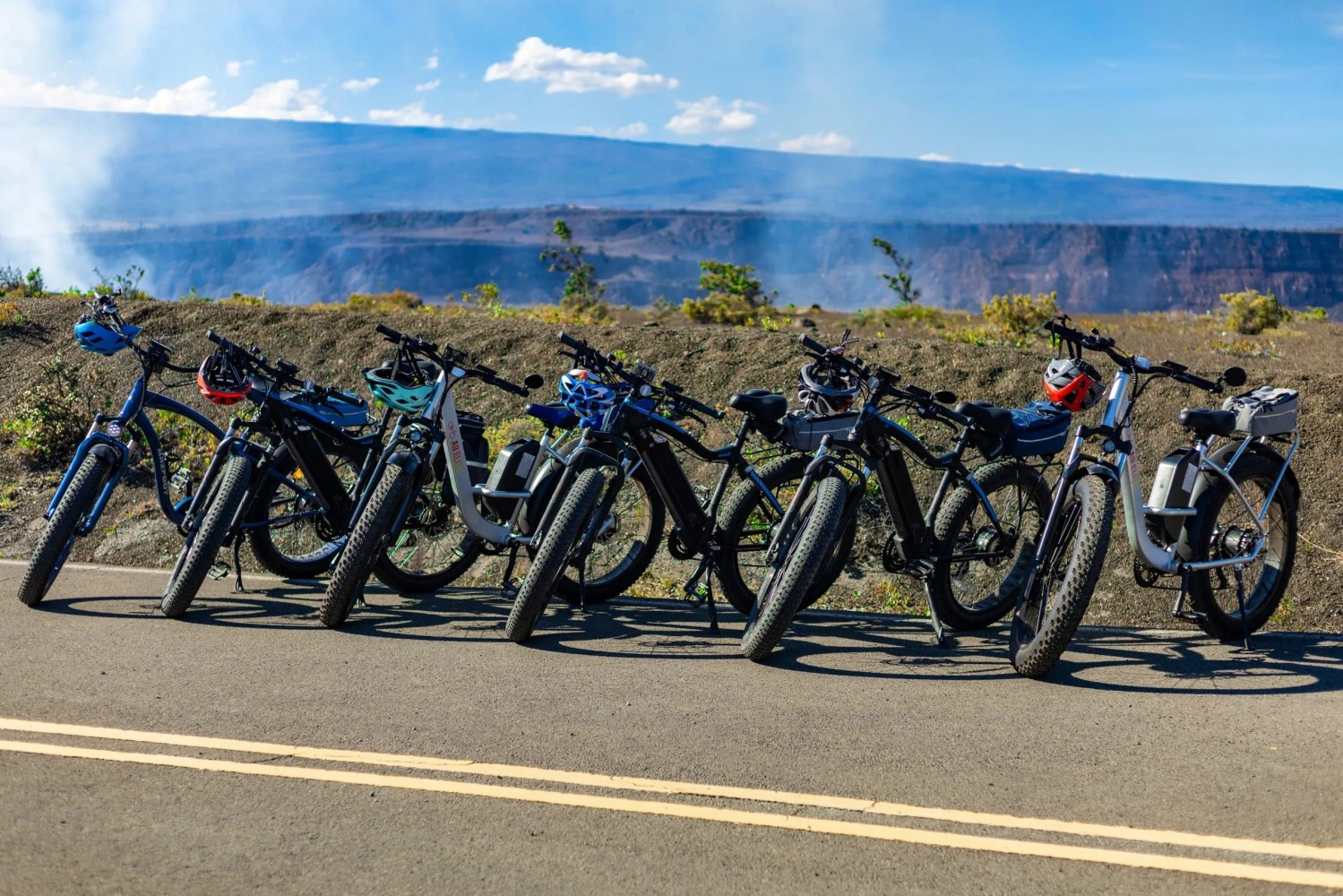 Seven electric bikes lined up on a road with helmets, overlooking a scenic canyon.