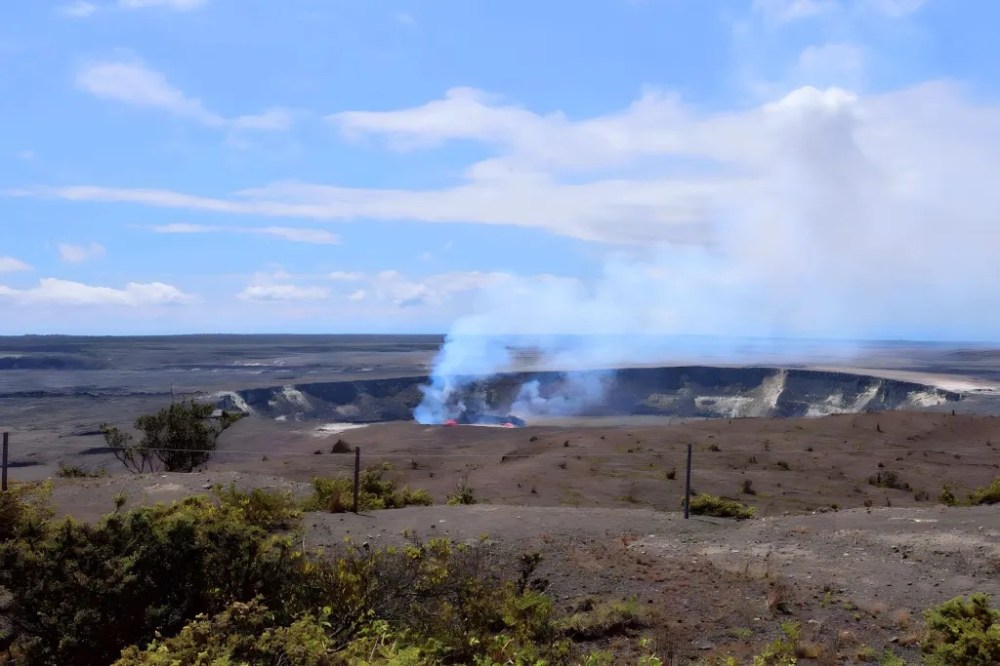 Volcanic crater with smoke rising under a clear blue sky, surrounded by barren landscape.