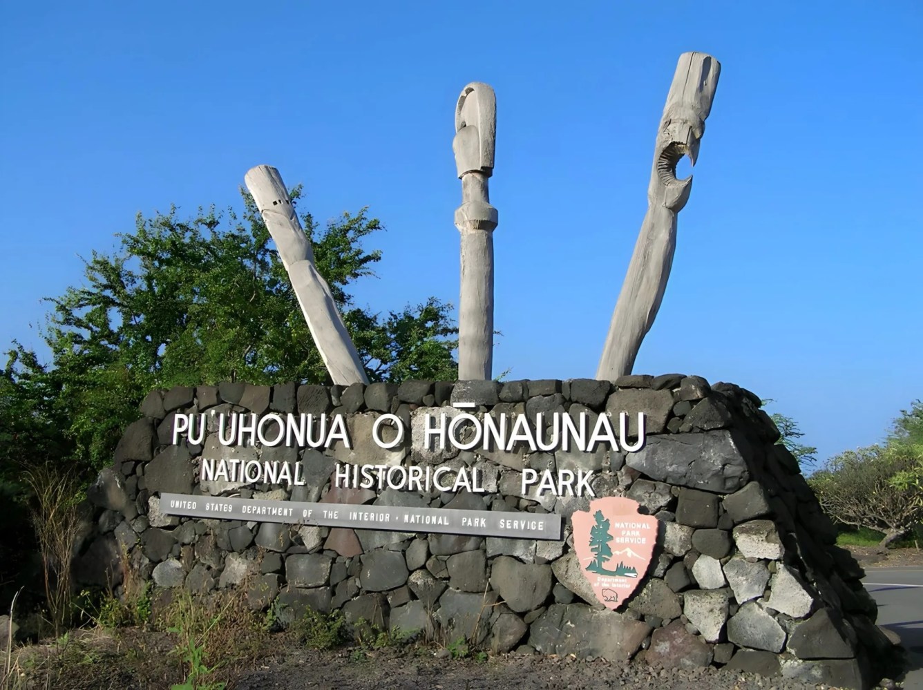 Sign for Pu'uhonua o Hōnaunau National Historical Park with stone base and wooden carvings.