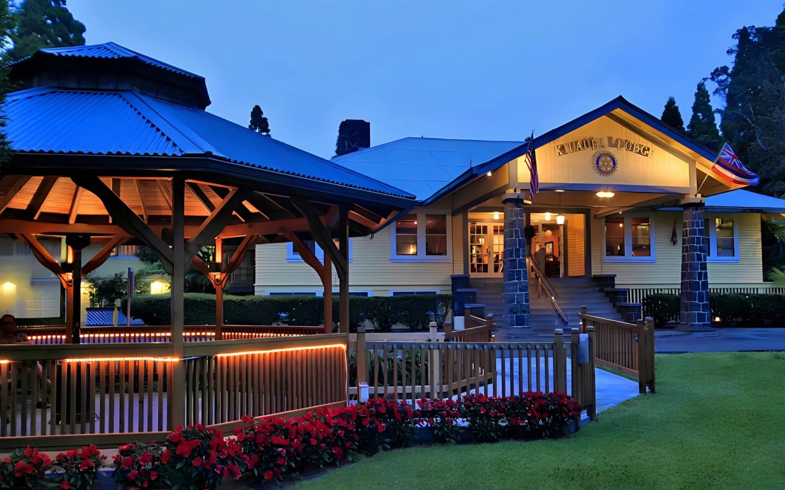 A lit gazebo and a yellow lodge building with flags, surrounded by flowers and trees at twilight.