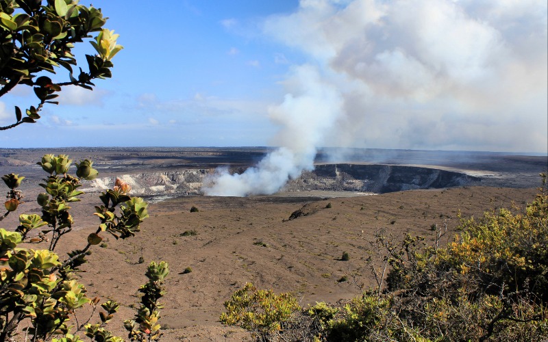 Volcano crater with smoke rising, surrounded by sparse foliage under a blue sky.