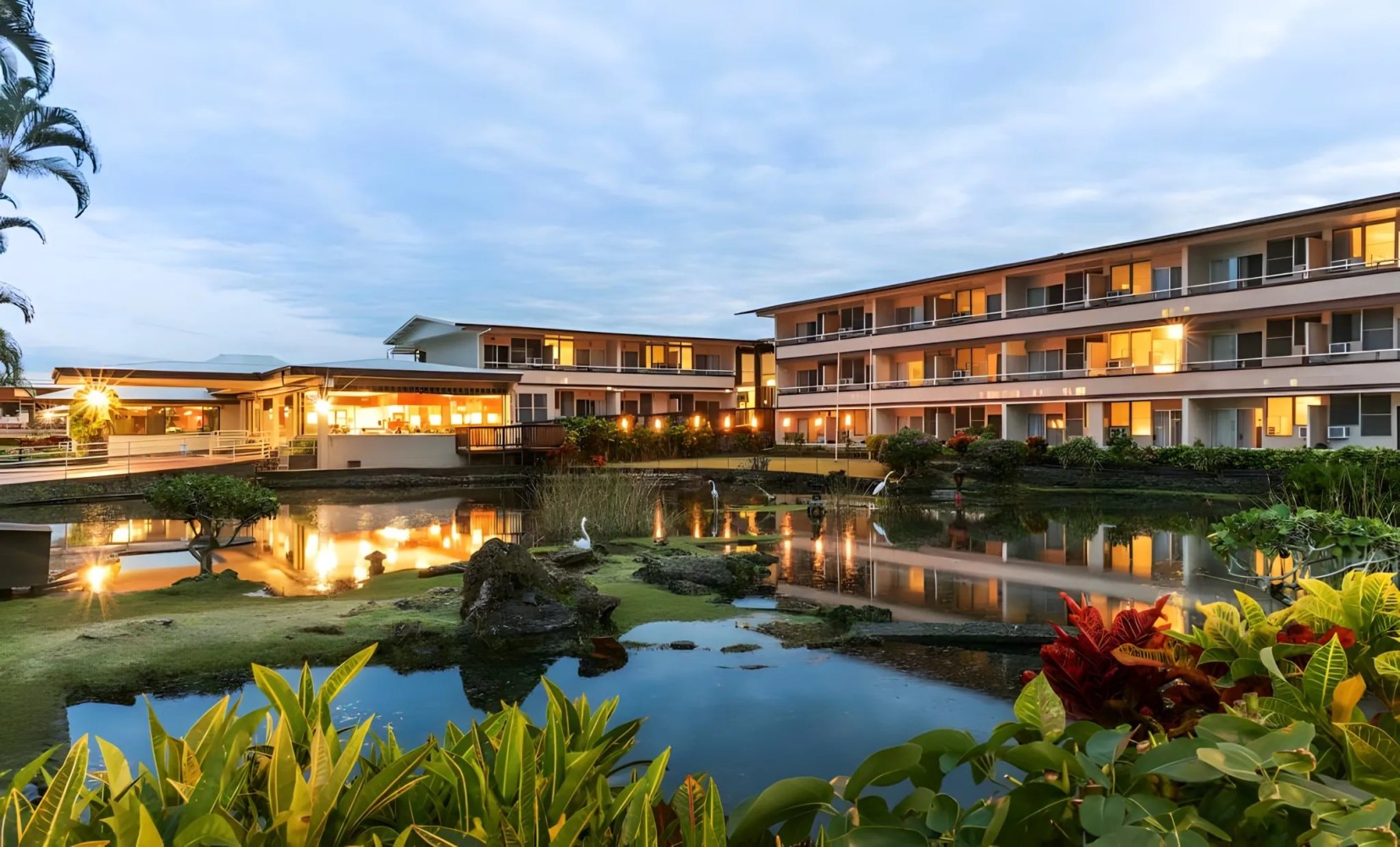 Exterior view of a hotel building with lit windows and a reflective pond in the foreground at dusk.