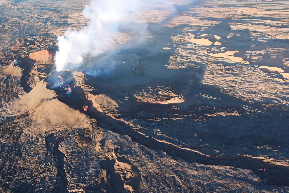 Aerial view of an erupting volcano with smoke and lava flow in a rugged landscape.