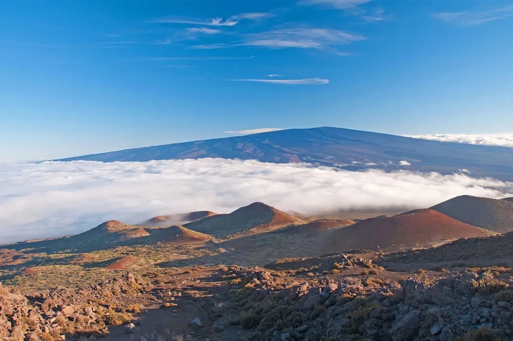 Mountain landscape with rocky terrain, blue sky, and clouds below a distant peak.