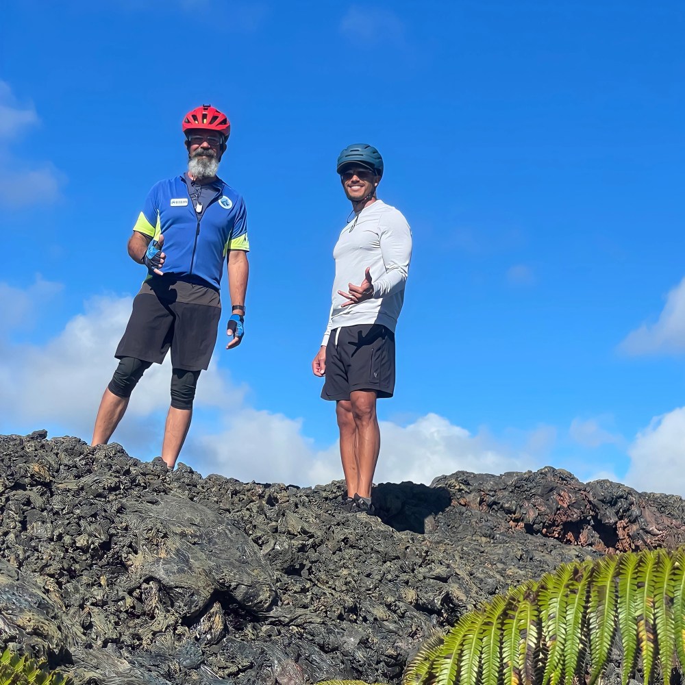 Two cyclists in helmets stand on rocky terrain under a clear blue sky.