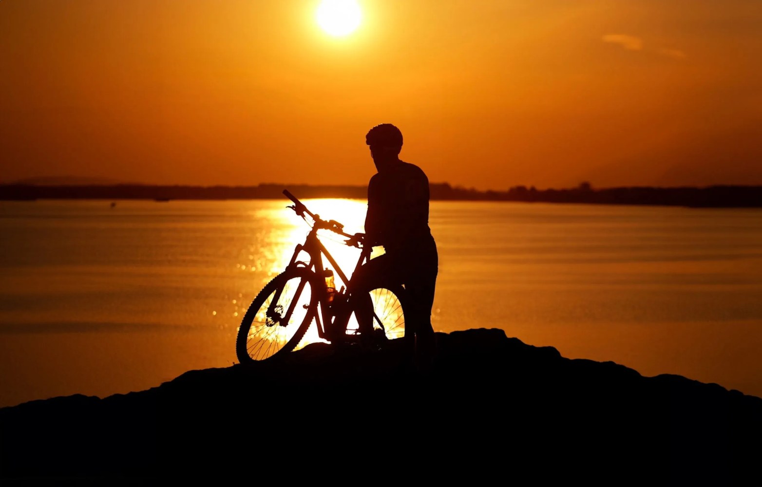 Silhouette of a person with a bicycle against an orange sunset over water.