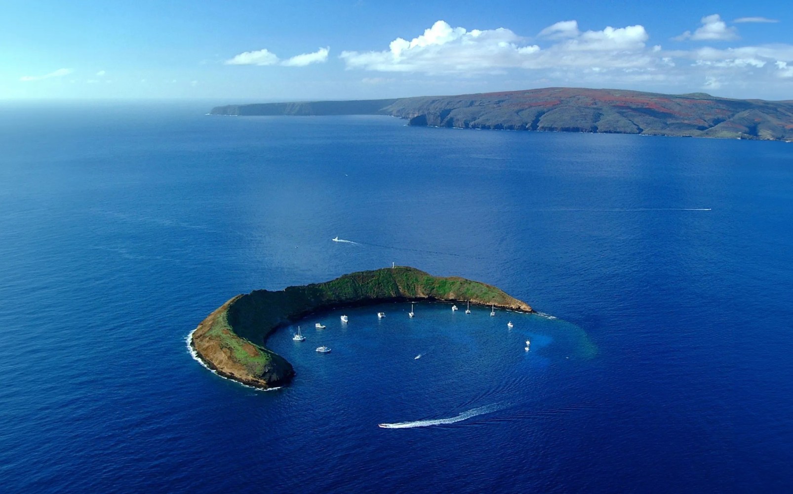 Aerial view of crescent-shaped island with boats in a blue ocean and distant land on the horizon.