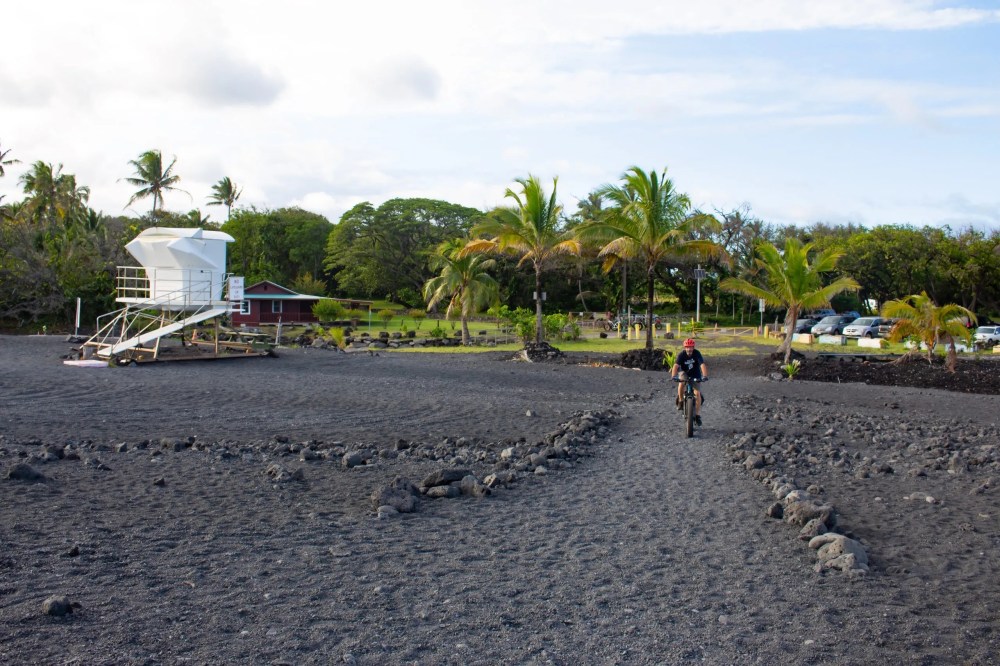 Person biking on a black sand beach near a lifeguard tower and palm trees.